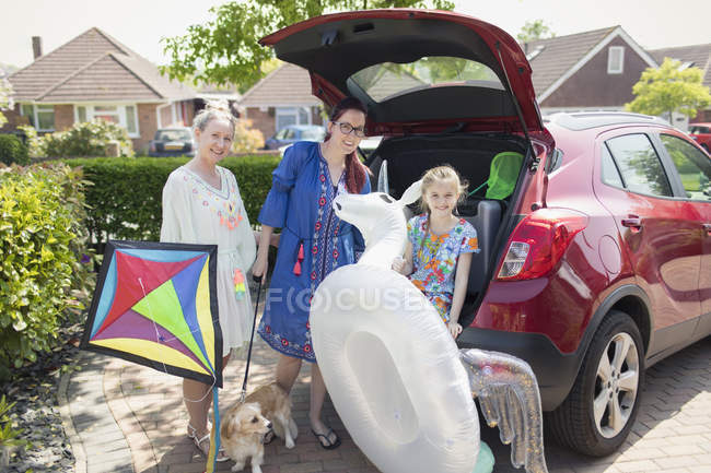Retrato lesbiana pareja e hija con cometa y unicornio inflable cargando coche en la calzada - foto de stock