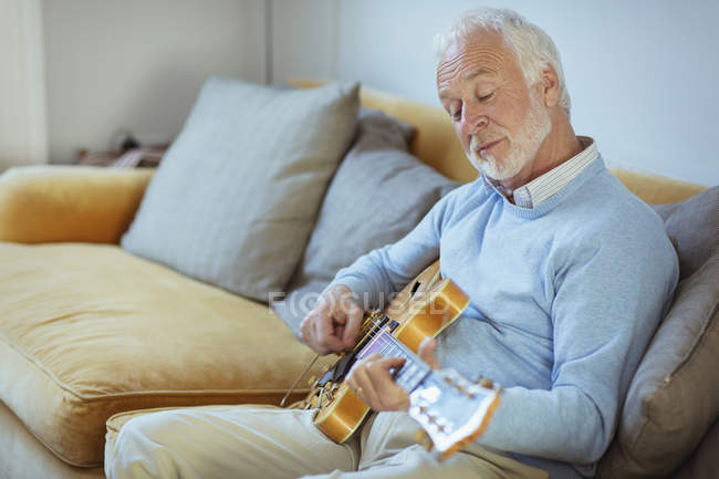Uomo anziano attivo che suona la chitarra sul divano del salotto — Foto stock