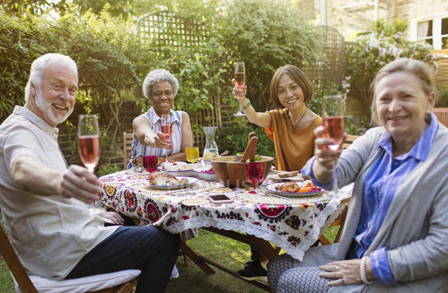 Retrato sorridente, confiante amigos seniores ativos beber vinho rosa e desfrutar de almoço na mesa do pátio — Fotografia de Stock