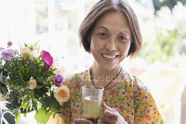 Retrato sorrindo, mulher sênior confiante bebendo chá ao lado de buquê de flores — Fotografia de Stock
