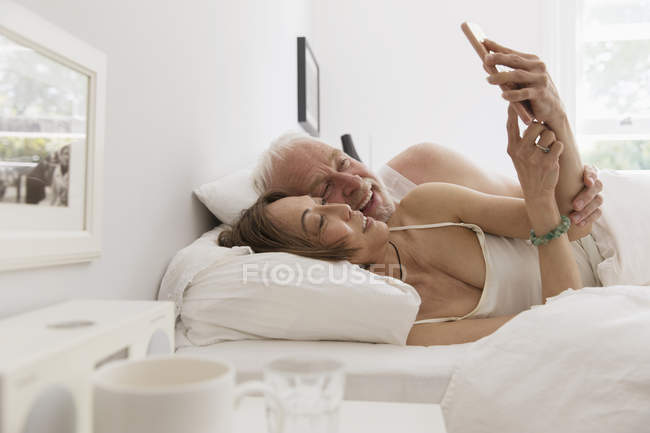 Affectueux couple de personnes âgées en utilisant un téléphone intelligent au lit — Photo de stock