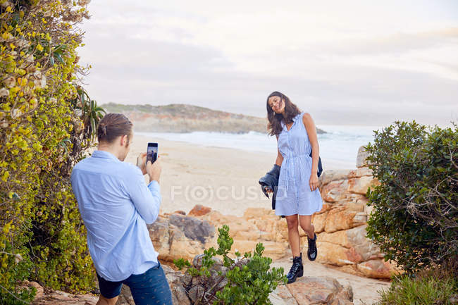 Young man with smart phone photographing girlfriend with ocean in background — Stock Photo