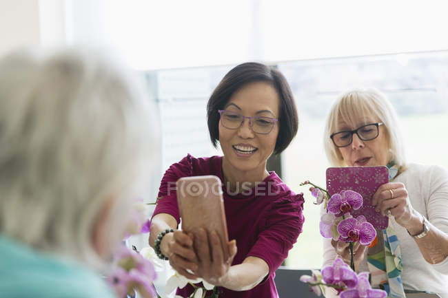 Femmes âgées actives avec appareil photo téléphone photographier des orchidées en classe d'organisation de fleurs — Photo de stock