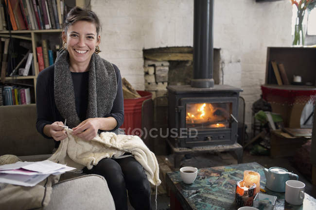 Retrato sonriente, mujer confiada tejiendo junto a la chimenea en la sala de estar - foto de stock