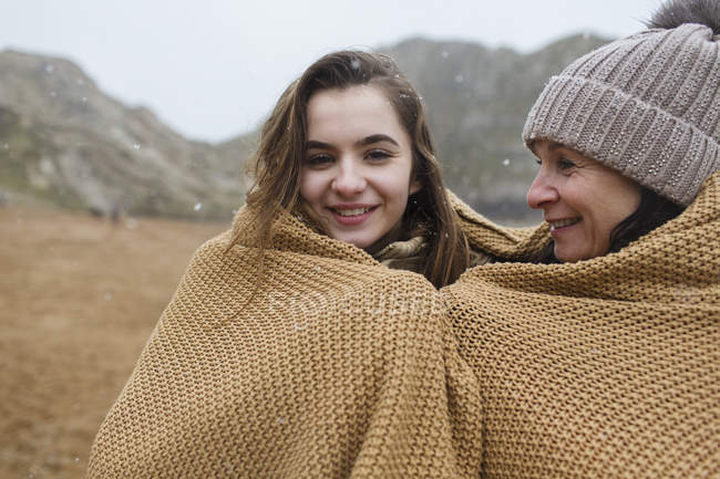 Retrato despreocupado madre e hija envueltas en una manta en la playa nevada de invierno - foto de stock