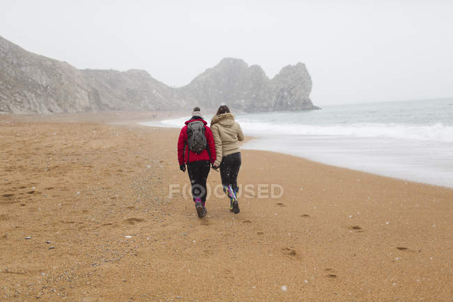 Madre e hija en ropa de abrigo caminando en la playa nevada del océano de invierno - foto de stock