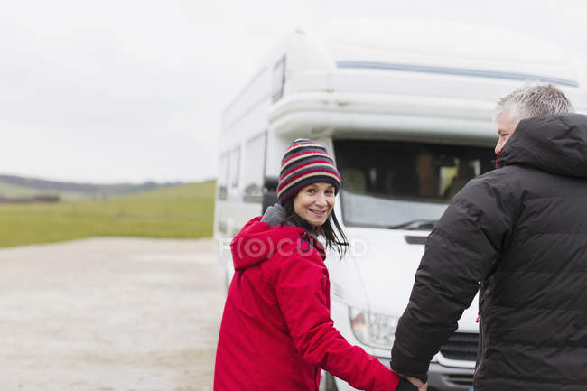 Портрет улыбающейся пары в теплой одежде, держащейся за руки возле дома на колесах — стоковое фото