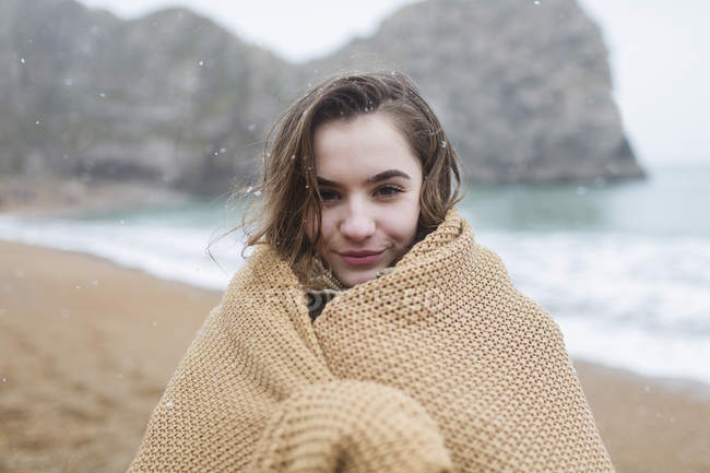 Portrait smiling teenage girl wrapped in blanket on snowy winter beach — Stock Photo