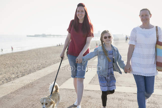 Casal lésbico afetuoso com filha e cachorro andando no calçadão ensolarado da praia — Fotografia de Stock