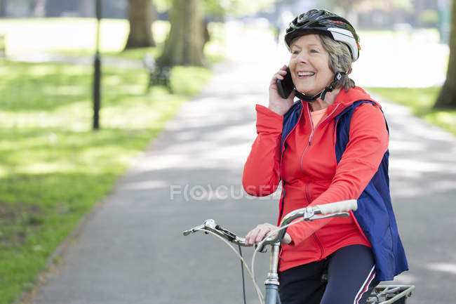 Mulher sênior ativa falando no telefone celular na bicicleta no parque — Fotografia de Stock
