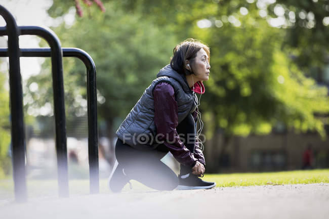 Active senior female runner with headphones tying shoe in park — Stock Photo