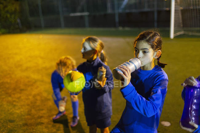 Girl soccer player taking a break, drinking water on field at night — Stock Photo
