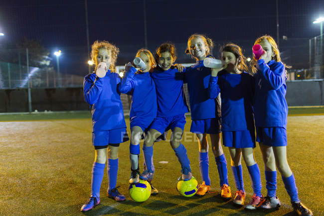 Ritratto fiducioso ragazze squadra di calcio bere acqua sul campo di notte — Foto stock