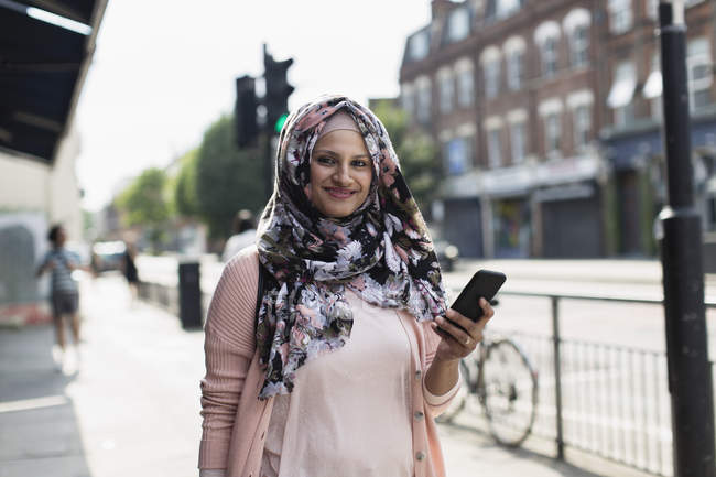 Retrato sonriente, mujer segura con teléfono inteligente usando hijab floral en la acera urbana - foto de stock