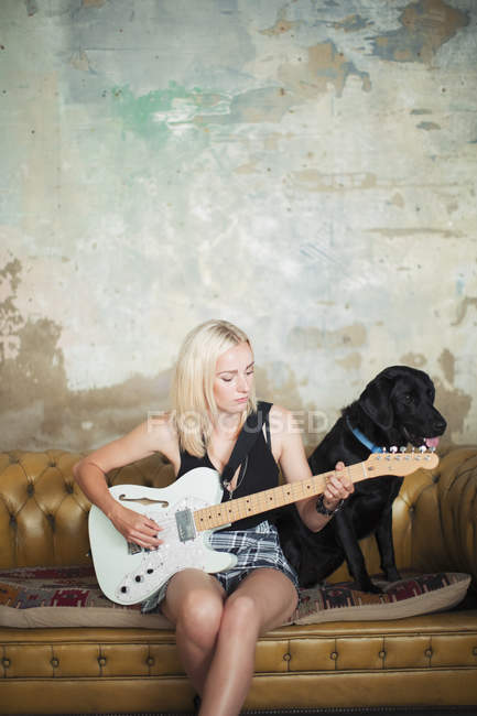Молода жінка з собакою грає на електричній гітарі на дивані — стокове фото