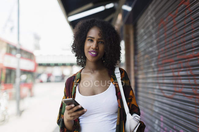 Retrato sonriente, joven confiada con teléfono inteligente en la acera urbana - foto de stock