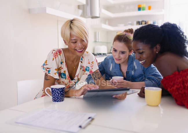 Молоді жінки друзі п'ють каву і використовують цифровий планшет за кухонним столом — стокове фото