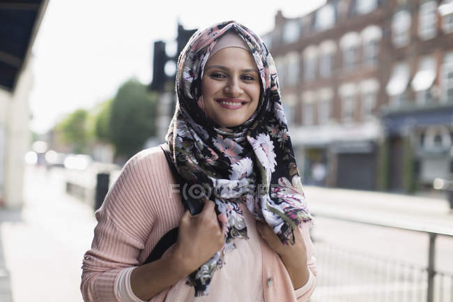 Retrato sonriente, mujer joven con hijab floral en la acera urbana - foto de stock