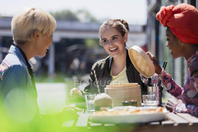 Jóvenes amigas comiendo almuerzo dim sum en la cafetería de la acera soleada - foto de stock