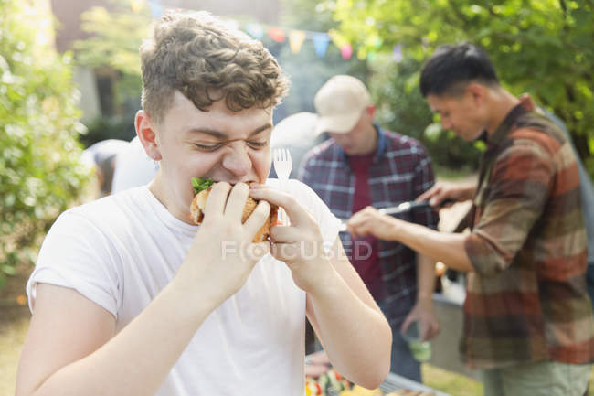 Голодний хлопчик-підліток їсть гамбургер на барбекю на задньому дворі — стокове фото