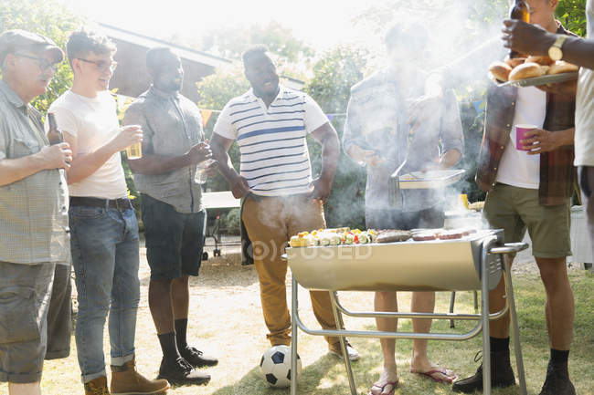 Мужчины пьют пиво и устраивают барбекю на солнечном летнем заднем дворе — стоковое фото