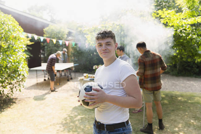 Retrato confiado adolescente con pelota de fútbol, disfrutando de barbacoa patio trasero - foto de stock