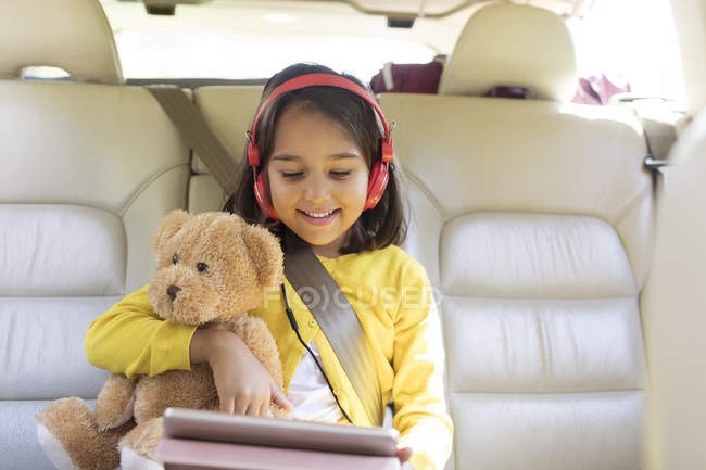 Fille souriante avec ours en peluche en utilisant une tablette numérique avec écouteurs sur le siège arrière de la voiture — Photo de stock