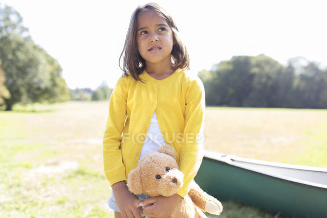 Menina com ursinho de pelúcia em campo ensolarado com canoa — Fotografia de Stock