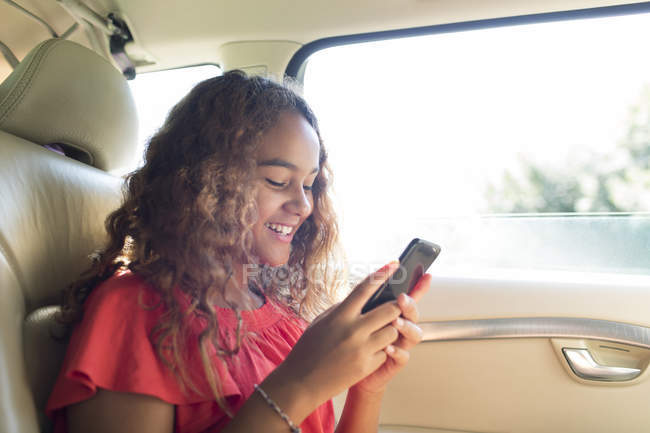 Sonriente chica adolescente usando el teléfono inteligente en el asiento trasero del coche - foto de stock