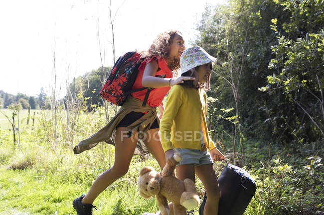 Sisters camping, carrying suitcase and teddy bear in sunny field — Stock Photo