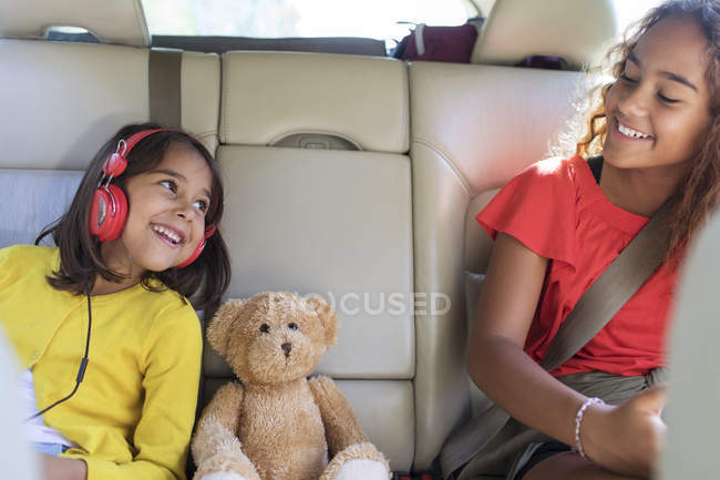 Happy sisters and teddy bear riding in back seat of car — Stock Photo