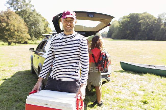 Porträt lächelnder Mann mit Campingkühltruhe, Auto in sonnigem Feld entladen — Stockfoto