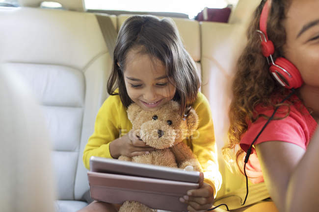 Jolie fille avec ours en peluche en utilisant une tablette numérique sur le siège arrière de la voiture — Photo de stock