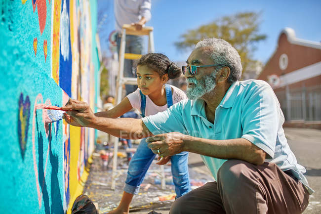 Avô e neta voluntários pintando mural vibrante na parede urbana ensolarada — Fotografia de Stock