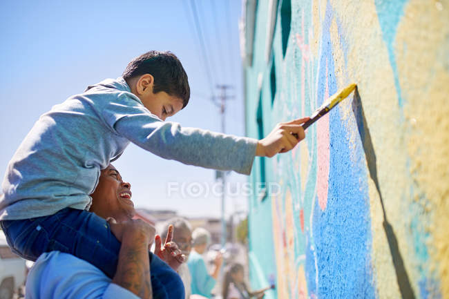 Padre e hijo voluntarios pintando mural en pared soleada - foto de stock