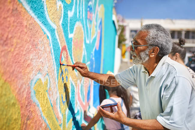 Senior male volunteer painting vibrant mural on sunny wall — Stock Photo