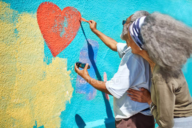 Senior couple painting heart-shape mural on sunny wall — Stock Photo