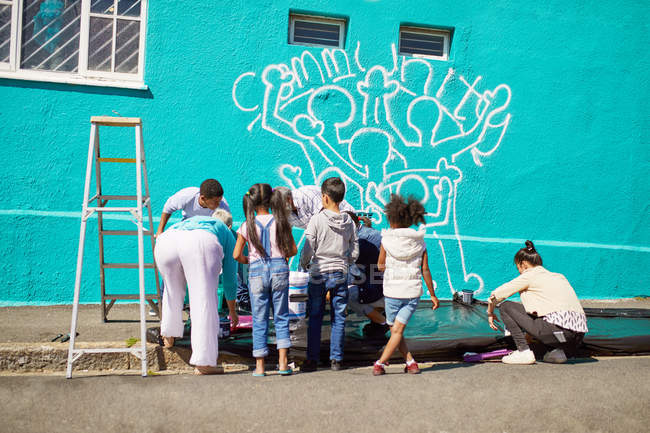 Enfants volontaires peignant une murale communautaire sur un mur ensoleillé — Photo de stock
