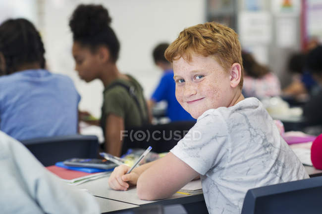 Портрет улыбающегося, уверенного ученика младшей школы, занимающегося за столом в классе — стоковое фото