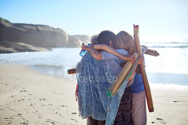 Mujeres amigas con colchonetas de yoga abrazándose en la playa soleada durante el retiro de yoga - foto de stock