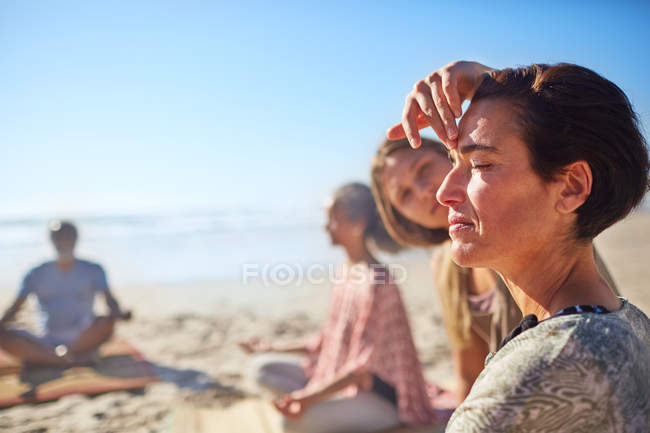 Instructor de yoga tocando el tercer ojo de la mujer meditando en la playa soleada durante el retiro de yoga - foto de stock