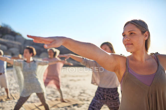 Mujer segura practicando la pose guerrera 2 en la playa soleada durante el retiro de yoga - foto de stock