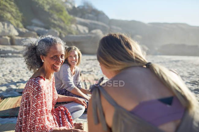 Mujeres sonrientes amigas hablando en la soleada playa durante el retiro de yoga - foto de stock