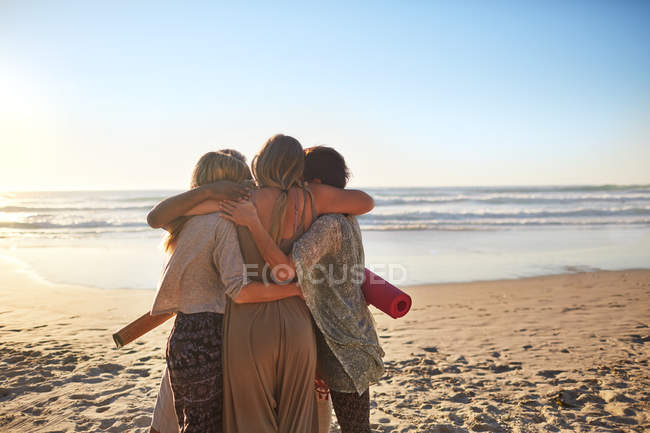 Amigas con colchonetas de yoga abrazándose en la soleada playa - foto de stock