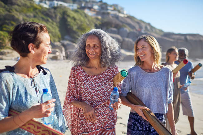 Mujeres amigas con colchonetas de yoga hablando en la playa soleada durante el retiro de yoga - foto de stock
