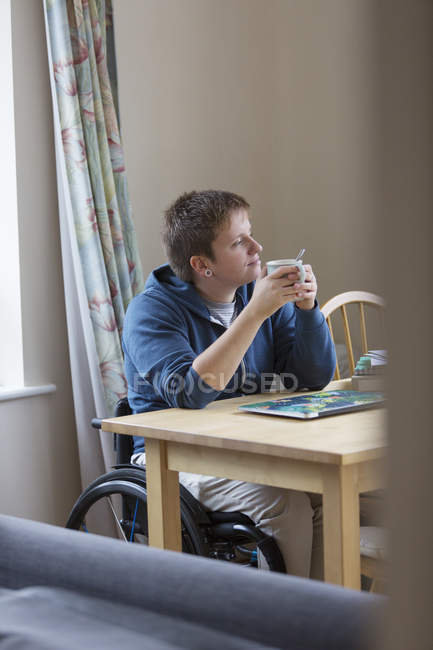 Задумчивая молодая женщина в инвалидной коляске пьет чай за обеденным столом — стоковое фото