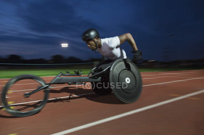 Determinato giovane atleta paraplegico maschile che accelera lungo la pista sportiva in gara su sedia a rotelle di notte — Foto stock