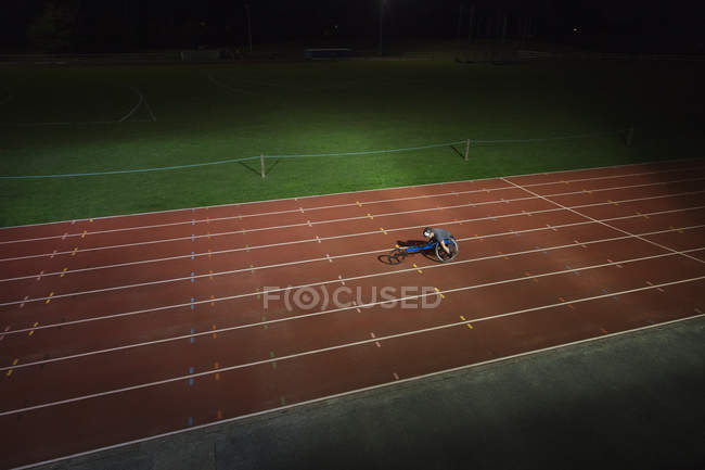 Atleta parapléjico exceso de velocidad a lo largo de la pista deportiva en la carrera en silla de ruedas por la noche - foto de stock