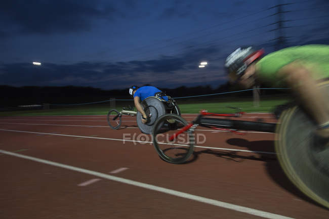 Atletas parapléjicos corriendo a lo largo de pista deportiva en carrera en silla de ruedas por la noche - foto de stock