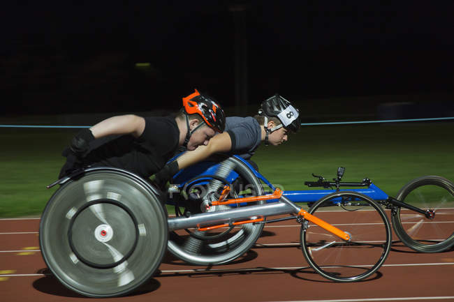 Atletas parapléjicos corriendo a lo largo de pista deportiva durante carrera en silla de ruedas por la noche - foto de stock